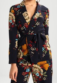 Blazer avvolgente in blu navy con maniche lunghe, caratterizzato da una cintura in vita e motivi floreali vivaci in arancione, rosso e blu su tessuto strutturato.