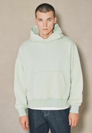 Young man with short hair wearing light green hoodie and dark jeans, standing against plain beige background, facing forward.