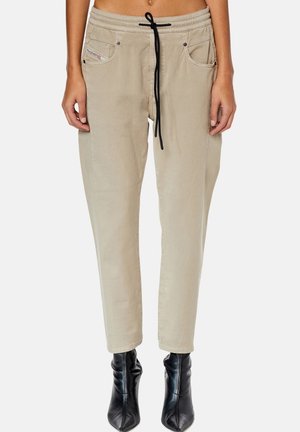 Beige casual broek met een zwarte trekkoord in de taille en voorzakken, gedragen met zwarte enkellaarzen door een staand persoon tegen een effen achtergrond.