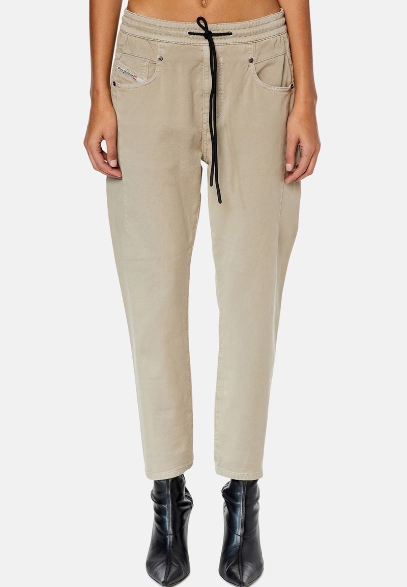 Pantalon beige décontracté avec taille à cordon noir et poches avant, porté avec des bottines noires sur une personne debout devant un fond uni.
