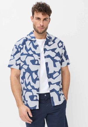 Mann trägt ein kurzärmeliges Hemd mit abstraktem blau-weißem Muster über einem weißen T-Shirt und steht vor einem schlichten hellen Hintergrund.