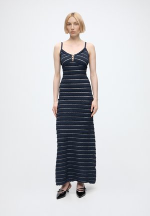 Frau mit kurzen blonden Haaren trägt ein dunkelblaues ärmelloses Maxikleid mit horizontalen Streifen und schwarze spitze Schuhe mit Riemen.