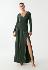Dlhé, hlbokozelené šaty s dlhými rukávmi a výstrihom do V. Obsahujú riasenie v oblasti pása a rozparok až po stehno. Hladká látka s jemným leskom.