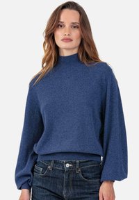 Pull en maille bleue avec un col montant et des manches longues et volumineuses. Ourlet et poignets côtelés. Porté avec un jean en denim foncé. Texture douce.