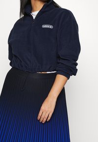 Kvinna iklädd en kort navy Adidas fleece med en vit logotyp och en högmidjad svart och blå veckad kjol, vänster arm avslappnad.