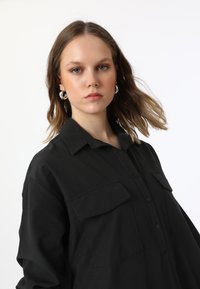 Chemise noire à boutons, en coton, présentant deux poches avant, un col, et une coupe décontractée avec une texture lisse.