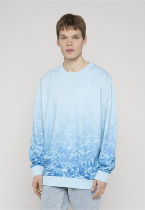 Ung mann iført en lys blå genser med et vannbølge-mønster, stående foran en ensfarget lys grå bakgrunn.