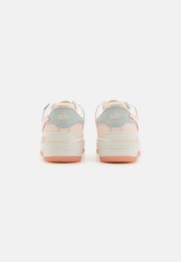 Sneakers Nike con una combinazione di colori bianco, rosa pallido e grigio. Presentano pannelli testurizzati, dettagli in cucito e una suola traslucida rosa.