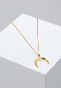 Collier en ton doré avec un pendentif courbé sur une chaîne délicate, présenté sur un piédestal géométrique blanc. Fini lisse et brillant.