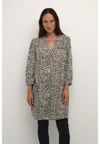 Robe tunique à pois noir et blanc avec décolleté en V, manches bouffantes longues et coupe décontractée, fabriquée en tissu léger. Portée sur un pantalon noir.