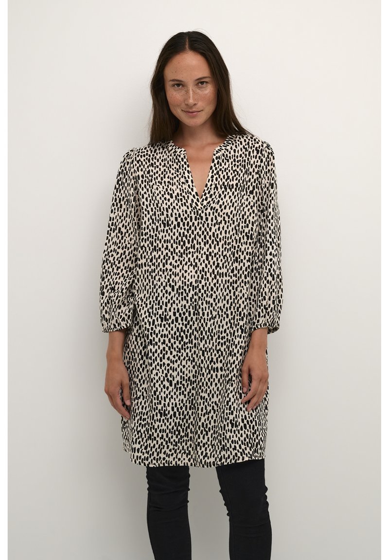 Robe tunique à pois noir et blanc avec décolleté en V, manches bouffantes longues et coupe décontractée, fabriquée en tissu léger. Portée sur un pantalon noir.
