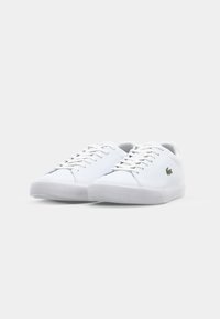 Adidași albi cu partea superioară din piele netedă, vârf rotund, șireturi plata și talpă din cauciuc texturată. Prezintă un mic logo verde de crocodil pe lateral.