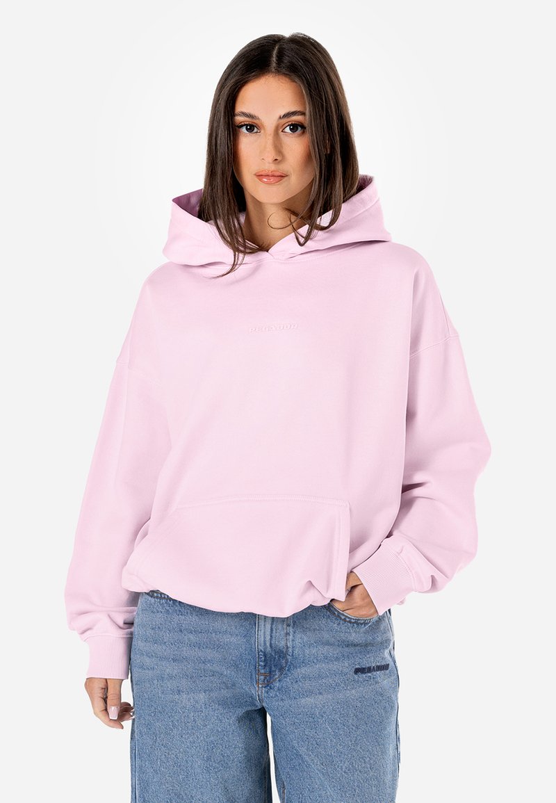 Hoodie oversized em rosa, com um bolso frontal, punhos canelados e capuz com cordão. Tecido suave; marcação discreta no peito. Combinado com jeans azuis.