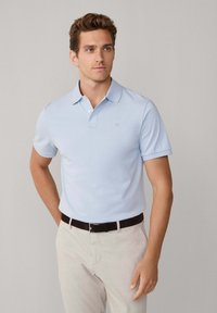 Lichtblauw poloshirt met kraag en korte mouwen, met een klein embleem op de borst, gecombineerd met beige gestructureerde broek en een bruine riem.