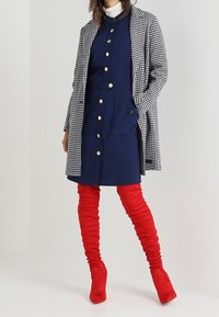 Žena v námořnických šatech na knoflíky, černo-bílém kostkovaném kabátu a červených semišových kozačk�ách po stehna stojící na šedém pozadí.