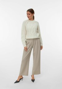 Vrouw die staat en naar beneden kijkt, gekleed in een witte kabelgebreide trui, beige wijde broek en zwarte kittenheels met stippen.