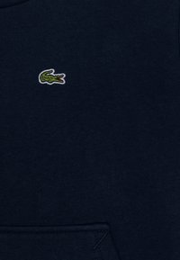 Tamsiai mėlynas džemperis su mažu žaliu krokodilo logotipu, pagamintas iš lygaus audinio, su priekiu kišene ir ribotais rankogaliais.