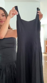 Kvinne med langt hår holder en ermeløs svart plisserte kjole med firkantet halsringning i et lyst rom med tregulv.