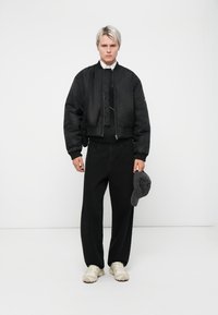 Chaqueta bomber negra con puños elásticos, superpuesta a un suéter negro. Pantalones anchos negros, zapatillas de color claro y gorra gris en la mano.