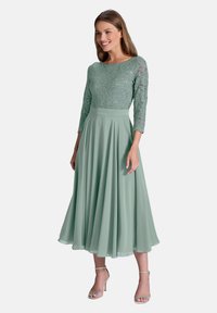 Robe vert menthe avec un corsage en dentelle, des manches trois-quarts et une jupe plissée fluide. Éléments de sequins discrets et taille cintrée.