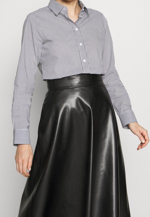 Mujer con falda negra de cuero sintético de talle alto y camisa blanca de manga larga con finas rayas negras verticales.