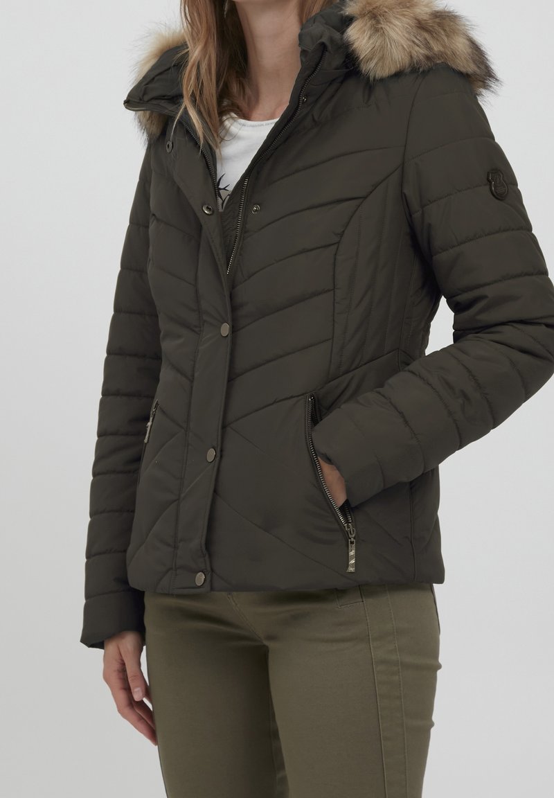 Veste matelassée vert foncé avec une capuche bordée de fausse fourrure, design matelassé, poches latérales zippées et boutons-pression. Portée sur un haut blanc.