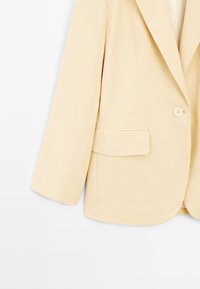 Beżowa marynarka na jeden guzik z klapami z wycięciem i kieszenią z patką, prezentowana na białym tle.