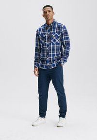 Niebieska koszula w kratę z dwiema kieszeniami na piersi, zestawiona z granatowymi spodniami i białymi sneakersami. Charakteryzuje się wzorem w kratę w kontrastowych kolorach.