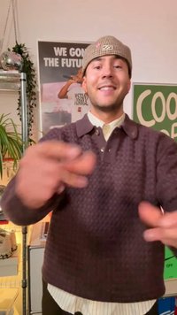 Bruine gebreide trui met textuur en een kraag, gedragen over een lichtgekleurde blouse. Handen maken gebaren naar de kijker in een informele setting.