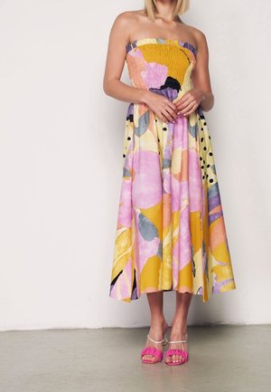Mujer con vestido midi sin tirantes, estampado floral colorido, corpiño fruncido y sandalias de tacón rosas, de pie contra una pared lisa.