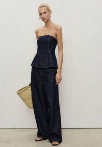 Denim peplum top a široké džíny v tmavě modré barvě. Oblečení má kontrastní švy a je doplněno tkanou slaměnou taškou.