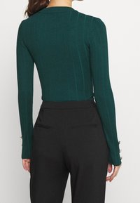 Dunkelgrünes, geripptes Langarmoberteil mit einer taillierten Passform. Schwarze, hochgeschnittene Hose mit minimalistischen Taschendetails auf der Rückseite.