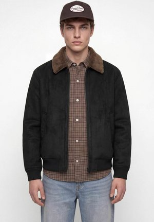 Jeune homme portant une veste bomber noire avec un col en fourrure marron, une chemise à carreaux marron, un jean bleu clair et une casquette noire, regardant vers l'avant.