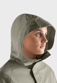 Chaqueta de lluvia de color verde oliva claro con capucha, que presenta una textura suave y a prueba de agua, cierre a presión y detalles de costura visibles.