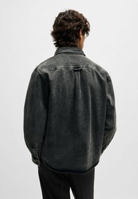 Persona con capelli ricci che indossa una giacca di denim nera, larga e sbiadita, e pantaloni neri, di spalle contro uno sfondo chiaro e semplice.