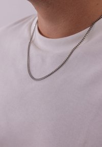 Zilveren ketting met een dunne, gevlochten ontwerp, gedragen boven een eenvoudig wit shirt. Het oppervlak weerkaatst het licht subtiel.