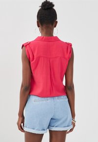 Blouse rose sans manches avec détails aux épaules et pli au dos, associée à un short en denim bleu clair. Tissu simple, coupe décontractée.
