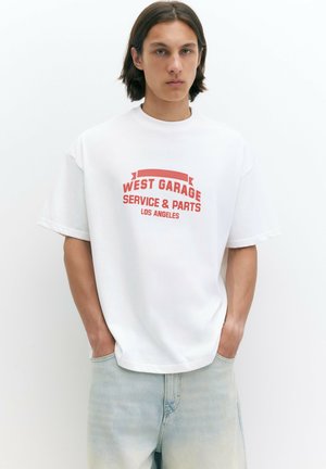 Ung mand med skulderlangt mørkt hår iført hvid T-shirt med rød tekst "West Garage Service & Parts Los Angeles" og lyseblå jeans.