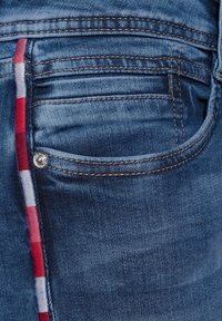 Jean en denim bleu avec une texture légèrement délavée, mettant en avant un motif à carreaux rouge et blanc visible à l'ouverture de la poche. Détail en rivet argenté.
