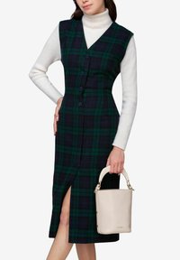 Grøn og marineblå plaidkjole med V-hals, knapfront og sideslids, båret over en hvid ribbet turtleneck. Holder en beige taske.