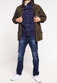 Olivově zelená bunda s kapucí přes tmavě modrou košili na knoflíky, doplněná modrými džíny a bílými teniskami. Látka působí hladce, volný střih.