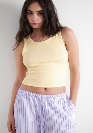 Młoda kobieta z długimi ciemnymi włosami, ubrana w bladożółty bezrękawnik i jasnofioletowe, w paski spodnie ze sznurkiem.
