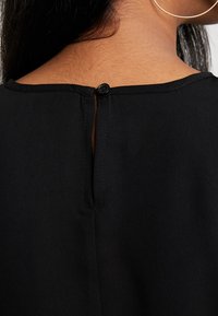 Gros plan sur le cou et le haut du dos d'une femme portant un haut noir fermé par un petit bouton noir au niveau de l'encolure, avec une boucle d'oreille en or visible.