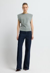 Donna che indossa un top grigio senza maniche con spalline, jeans a vita alta blu scuro e scarpe appuntite color borgogna, in piedi davanti a uno sfondo bianco.