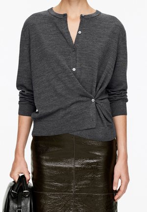 Femme portant un pull portefeuille gris foncé boutonné et une jupe en cuir noire brillante, tenant un sac à main noir dans la main gauche.