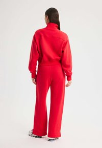 Rote Sweatshirt mit hohem Kragen und langen Ärmeln, kombiniert mit weit geschnittenen roten Hosen. Das Material wirkt weich, mit minimalen Nähten und einem lässigen Schnitt.