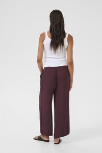 Woman with long brown hair wearing a white tank top, loose maroon pants, black slide sandals, standing facing away on a white background.