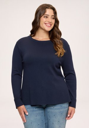 Kvinde med langt bølget hår iført en navyblå langærmet sweater og lyseblå jeans med små mørke pletter, smilende og kigger til siden.