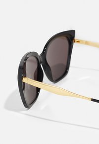 Lunettes de soleil noires au design distinctif en forme d'œil de chat, avec des verres foncés et des accents en métal doré sur les branches, gravées avec "vogue."