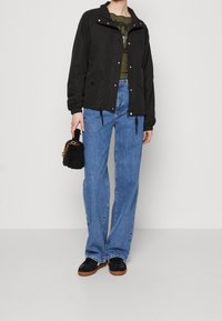 Person som bär en svart jacka, olivgrön topp, blå raka jeans, svarta sneakers med bruna sulor, och håller en svart texturerad handväska med guldkedja.